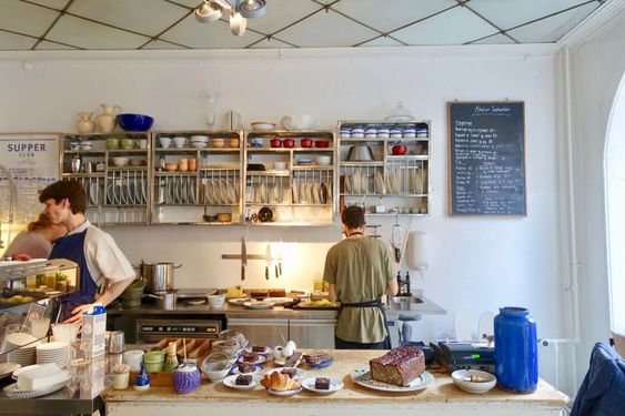 Kitchen and prep area of an over-the-counter cafe. Light environment with open-concept shelves featuring plate and mug varieties. Natural wood, white, and metal surfaces with notes of bright red, blue, lime, and pale yellow tableware. A chalboard menu hangs on the wall in the background, various pastries are nestled in the foreground. Cafe employees are keeping busy.